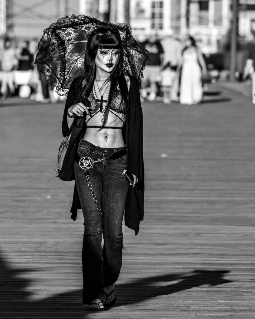 Black and white street photography of a woman in gothic fashion walking down a sunny boardwalk carrying a black lace parasol.